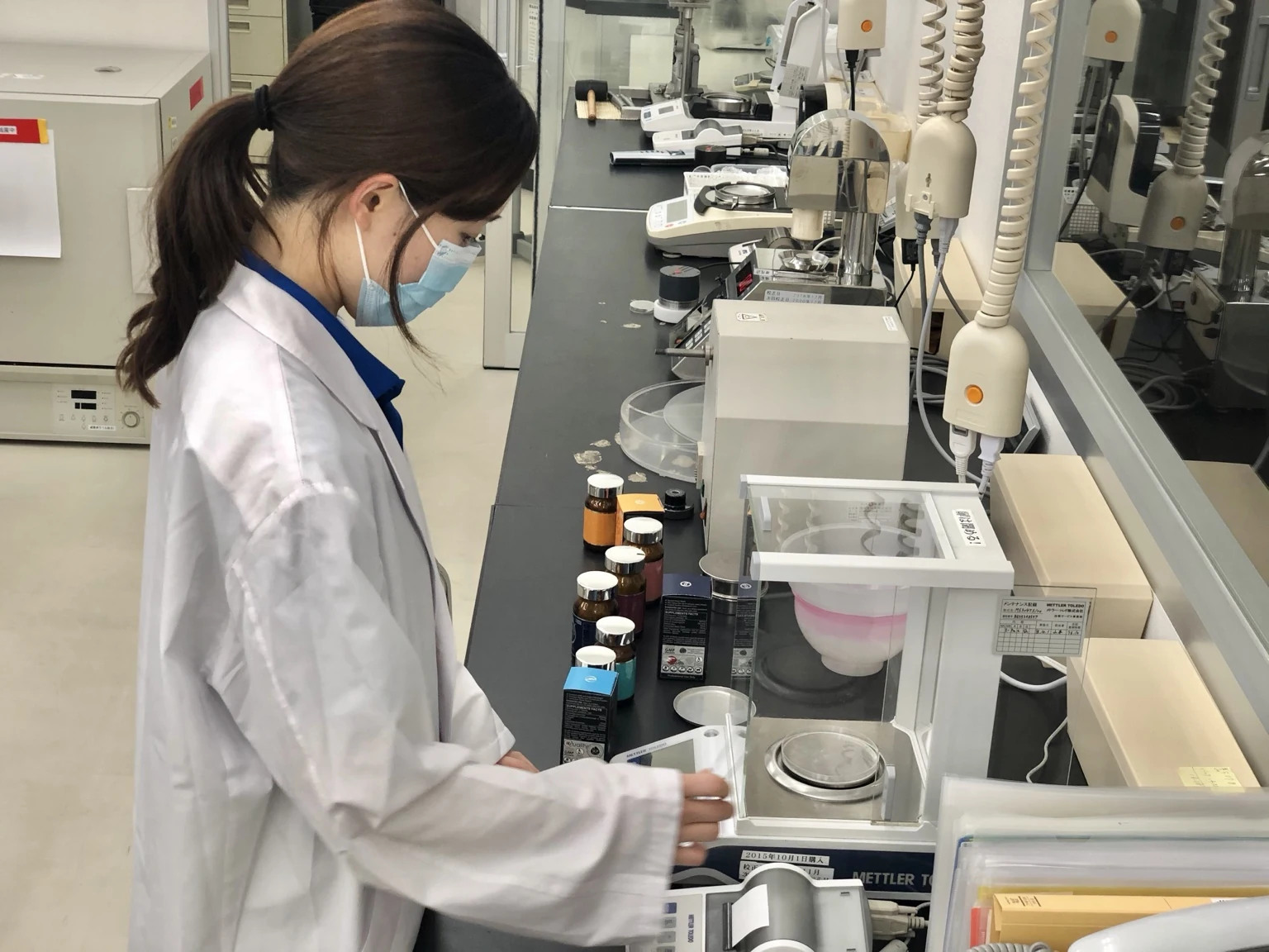 A laboratory specialist beside analytical balances and Yotsuba Japan product packaging at a Japanese manufacturing partner